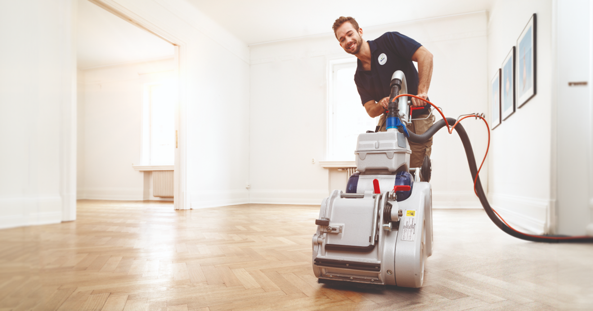 Smiling technician performing Wood Floor Sanding Prep on an oak herringbone parquet floor in Moorgate with a powerful belt sander.
